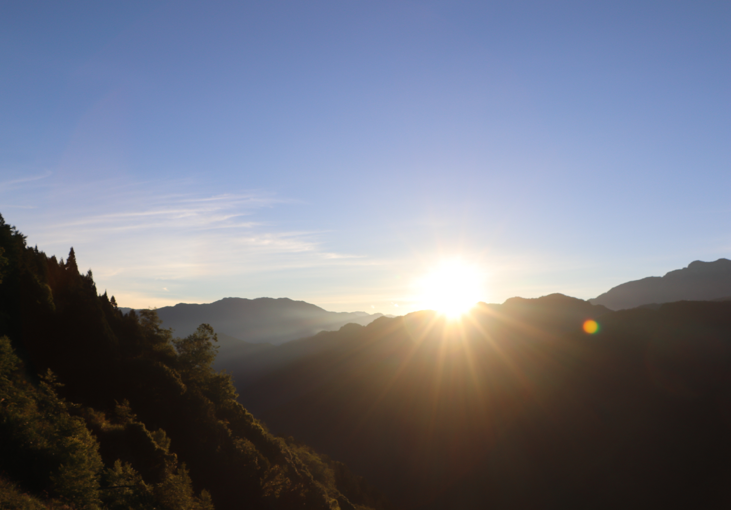 山谷晨曦景象，金色陽光從遠方山峰升起，照亮層疊的山脈輪廓。前景為深綠色森林，背景山巒籠罩在晨霧中，呈現寧靜祥和的自然風光。