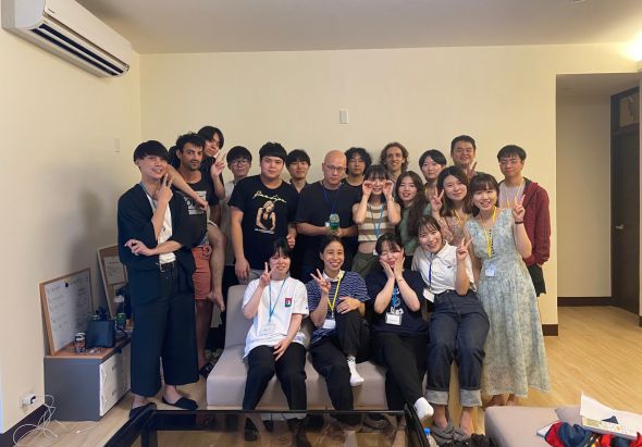 室內活動合照，約二十多位參與者穿著各式服裝，在米色牆壁前排成兩排。前排坐著，後排站著，眾人面帶笑容擺出歡樂姿勢。背景可見冷氣機、門框和木質地板，營造溫馨團聚氛圍。