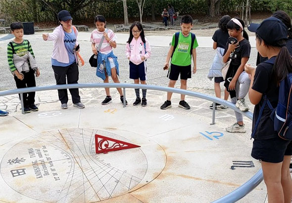 一群學童圍成一圈，站在校園空地上進行戶外教學活動。地面上繪有圓形圖案和紅色三角旗幟標記，老師正為學童講解指導，孩子們專注觀看和學習。