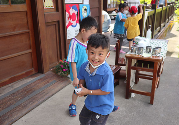 戶外活動場景中，一名穿著藍色polo衫的小男孩開心地笑著，手中拿著玩具。身後還有其他小朋友。木製建築物、紅色木門和擺放整齊的木製椅子構成古樸的傳統風格背景。
