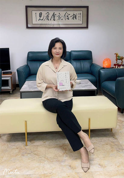 一位女性坐在淡黃色沙發上，穿著米色襯衫和黑色褲子，手持書籍。身後是綠色沙發、書架和牆上的中文書法作品。室內裝潢現代簡約，展現專業氛圍。