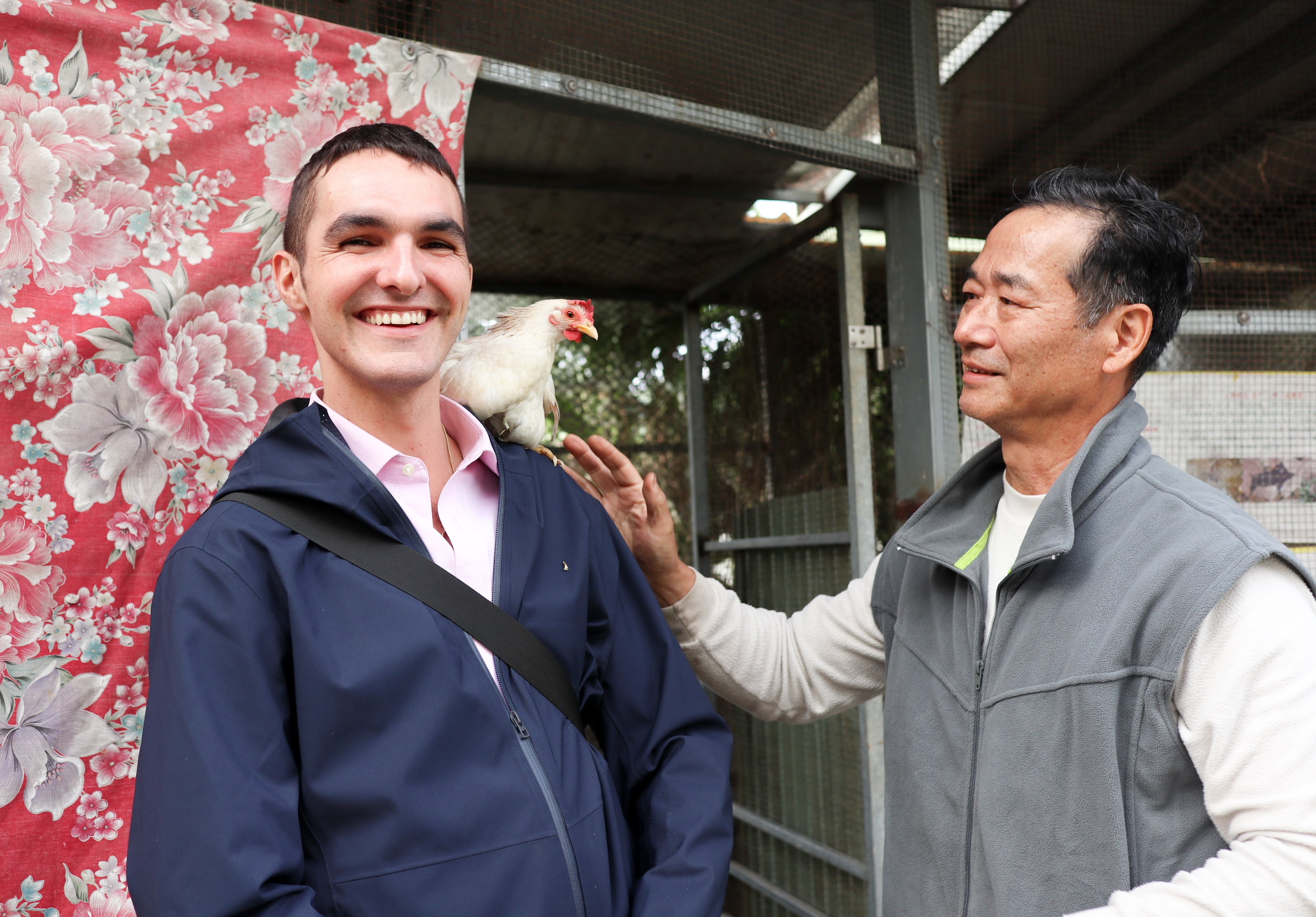 兩名男子在室外場景互動，左側男子穿著深藍色夾克，笑容燦爛，肩上站著一隻白色的雞。右側男子穿著灰色背心，正用手輕輕觸碰雞。背景有紅色花卉圖案布料及鐵製建築結構。