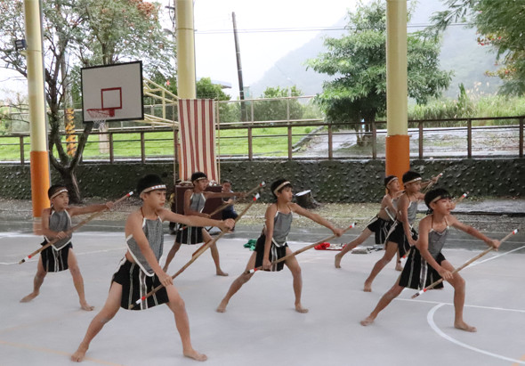 一群小學生穿著黑色運動服在室外籃球場進行體操表演，做出各種伸展和跨步動作。背景有籃球架、綠色圍籬和山景。觀眾坐在看台上觀看表演。