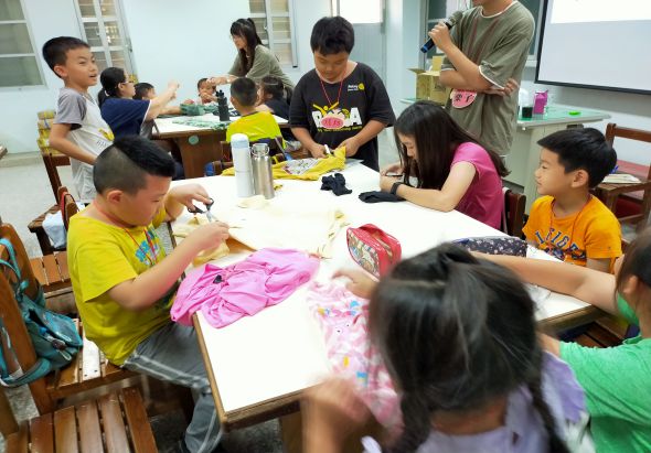 一間教室內，多名小學生圍坐在桌邊進行手工藝活動。前景一名穿黃色衣服的男童正在裝飾粉紅色布料，其他學生在各自的座位上進行類似創意工作。教室後方有學生站著，牆上有白板和儲物櫃，場景充滿互動學習的氛圍。