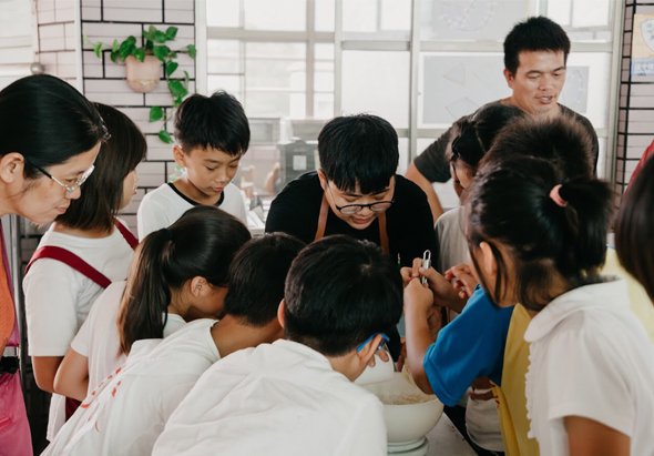 一群學生聚集在明亮教室內，圍繞著穿著黑色衣服戴眼鏡的教師，認真聆聽課程指導。窗戶背景採光充足，牆上掛著綠色植物裝飾。學生們專注於互動式學習活動。