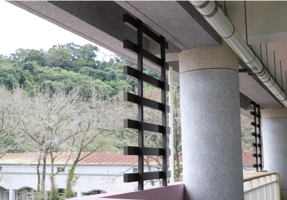 建築物遮陽棚下的黑色木製格柵屏風，旁邊豎立灰色圓形混凝土柱。背景可見綠樹覆蓋的山丘、紅色屋頂建築和白色護欄，展現現代建築與自然風景的結合。