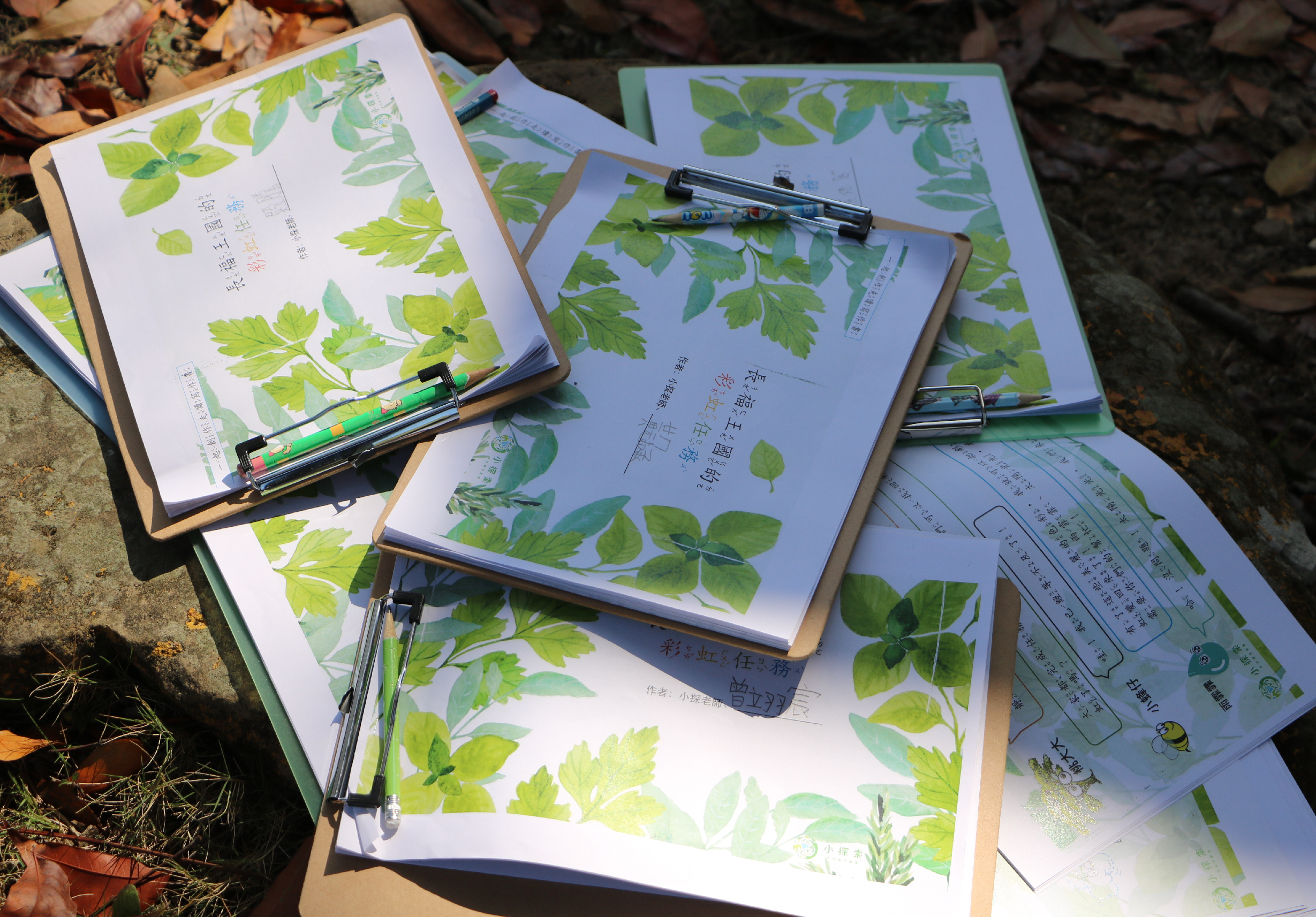 戶外進行植物學習活動，多本植物圖鑑展開放置於地面，書頁上有綠色葉片插圖與中文標註，配有彩色筆與測量工具，記錄植物特徵與分類資訊。