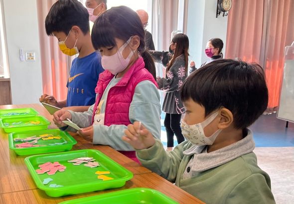 幼兒園教室內，數名戴著口罩的兒童圍坐在木製長桌前，正進行拼圖活動。桌上擺放綠色的拼圖盤，兒童專注地拼組圖片。背景有其他兒童站立活動，室內採光明亮。
