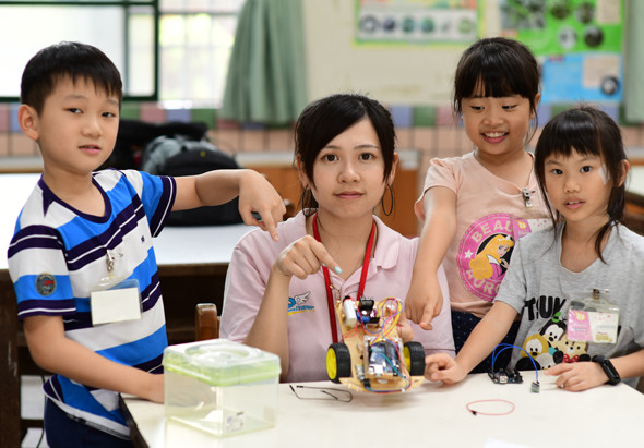 一名女性教師與四名小學生在教室內進行科學實驗課程。桌上擺放著實驗器材和裝置，學生們專注觀看並參與實驗過程，展現STEM教育的互動學習情景。
