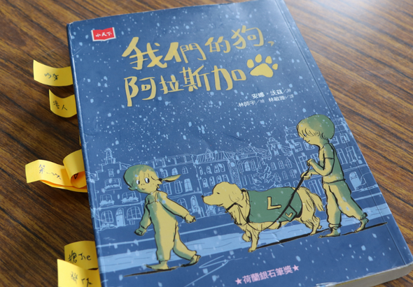 兒童繪本《我們的狗，阿拉斯加犬》封面，藍色夜空背景，黃色插圖展示兩個小孩與一隻狗互動的溫馨場景，書側邊有黃色書籤標籤。