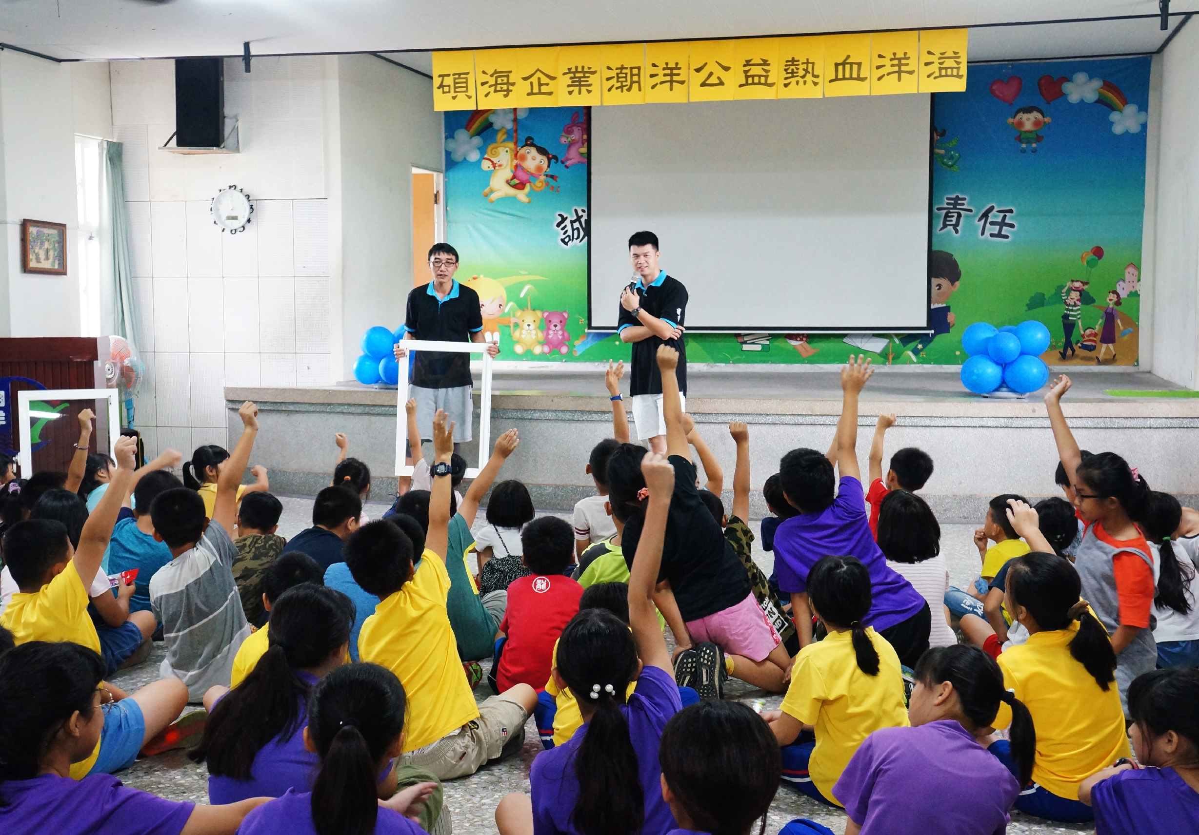 教室內舉辦公益活動，兩位講者在白板前向坐滿地板的小學生演講。學生穿著黃、紫色衣服，踴躍舉手。牆上掛著「石頭海企業潮洋公益熱血洋澳」標語及卡通布置。藍氣球點綴會場。