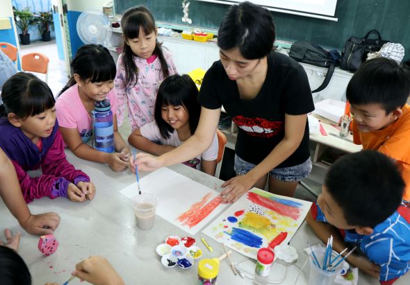 一位女性教師在教室中帶領六名小學生進行美術活動，學生們圍著白色工作台，桌上擺放著顏料盤、畫筆和調色板，學生們正認真地參與繪畫創作。