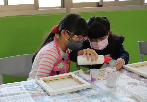 兩位戴著口罩的小學生在教室進行藝術創作課程。他們坐在鋪有報紙的桌子前，其中一名學生正在用白色容器倒液體到調色盤中。桌上放置著畫布、顏料和美術用具。綠色牆面和窗戶構成教室背景。