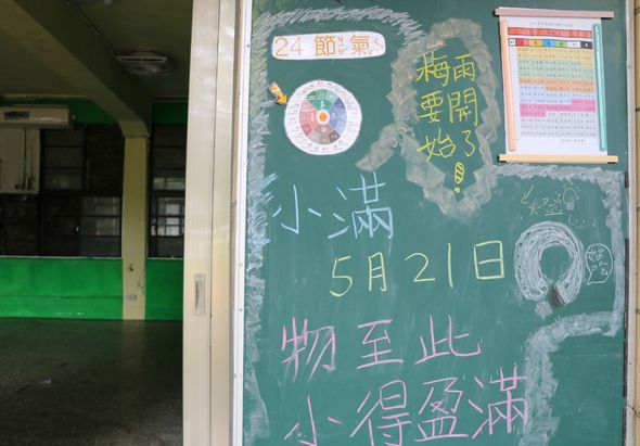 教室黑板上用粉筆寫著中文文字，包括「24節氣」、「5月21日」等內容，右側貼有彩色日曆表和教學海報，左方可見綠色課桌。