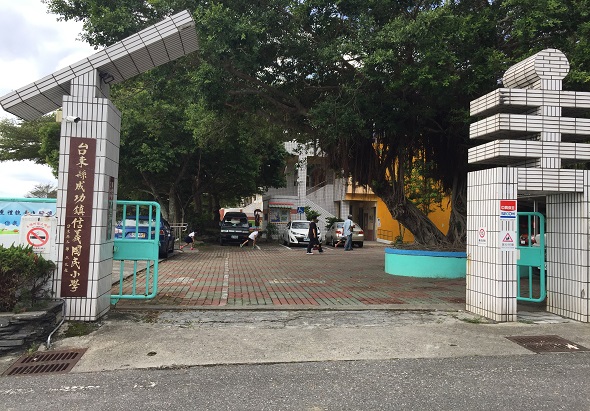 校園入口處，白色立柱式門樓兩側設置綠松石色售票亭。門樓內側為停車場，停有多輛車輛。背景為茂密大樹與建築物。地面為紅磚鋪裝。