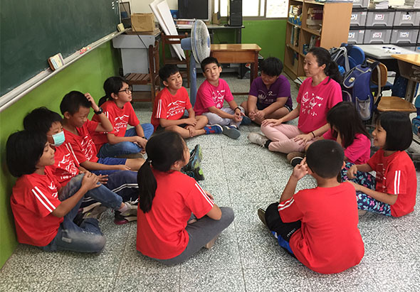 教室內，一群穿著紅色制服的學生圍坐成圓形，坐在地板上進行小組活動。他們專注地互動交流，背景可見綠色黑板、書架和教室設備。這是一堂集體學習或團隊建立的課堂活動。