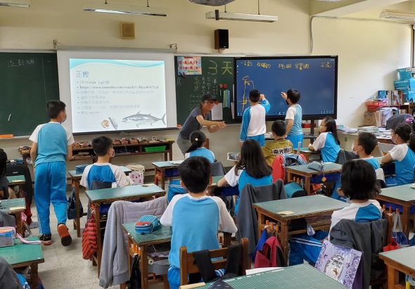 小學教室內，穿著藍色運動服的學生坐在課桌前聽講。講台上的教師指向投影螢幕，螢幕顯示魚類相關內容。黑板上有中文字跡。教室後方擺放各式教材和物品。這是一堂生物科學課程。