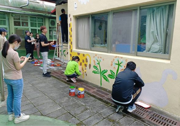 學童在校園戶外空地進行美術創作活動，數名學生蹲坐地面用顏料繪畫，另有學生站立觀看或等候，牆面上已有綠樹等彩繪圖案，展示集體創意的校園美化活動。
