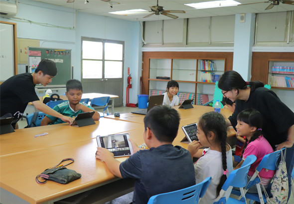 教室內，兩名成人教師與約七名小學生圍坐木製長桌，學生手持平板電腦進行課程學習。教室後方有書櫃、綠色黑板、滅火器和天花板風扇，呈現互動式教學情景。