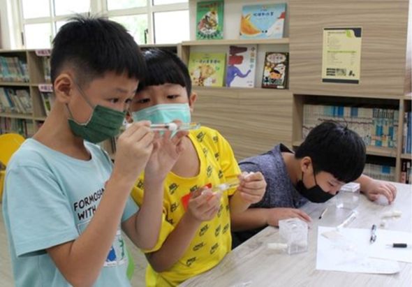 學校圖書館內，三名戴著口罩的學童圍坐在桌前進行科學實驗。左側男童和中央女童正在觀察實驗器材，右側男童低頭專注於實驗操作。桌上擺放著透明容器和實驗用具，身後為書架和教室布告欄。