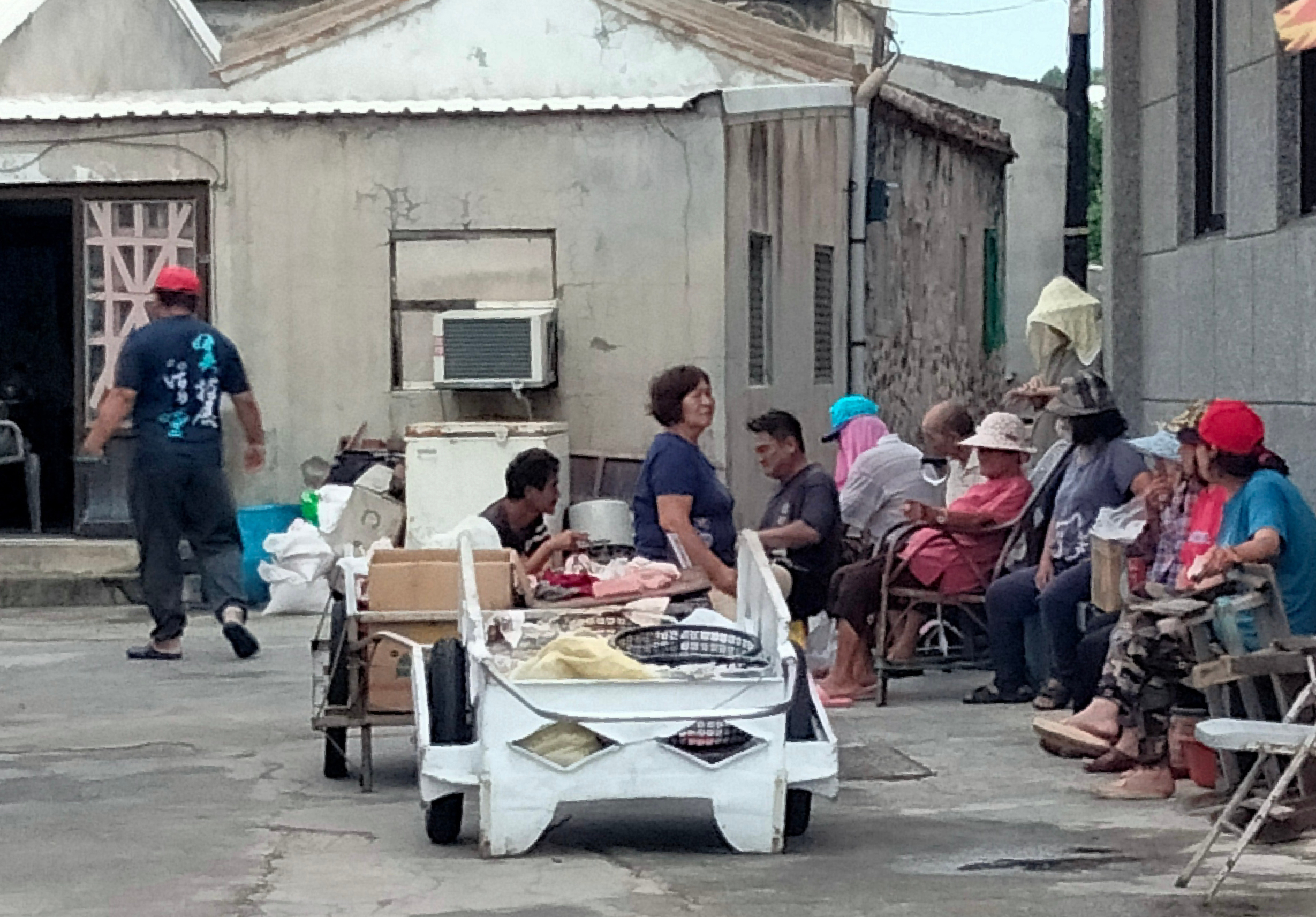 社區居民聚集在簡陋住宅區的空地上，圍坐成一圈參與社區活動。中央放置一台白色的移動式物資車，載有衣物和生活用品。多位穿著彩色衣服和帽子的民眾正在領取物資或交流互動，反映基層社區的互助精神。