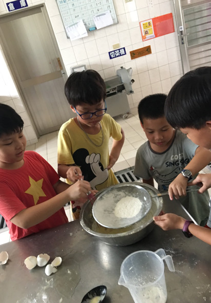 四名小學生在教室內進行烹飪活動。穿紅色星星衣服的男孩和穿黃色衣服的男孩共同持著大碗，展示白色麵糰或麵粉混合物。其他兩名學生在旁觀看協助。桌上放有食材和量勺，背景是教室布告欄。