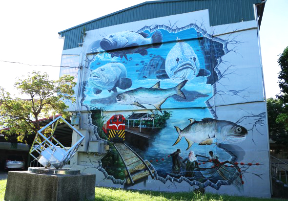 建築物外牆上繪製的大型海洋生物壁畫，描繪多種魚類、鯊魚及海洋場景。牆下方停放一輛紅色火車模型，周圍有綠色樹木。整幅壁畫以藍色調為主，展現豐富的海洋生態。