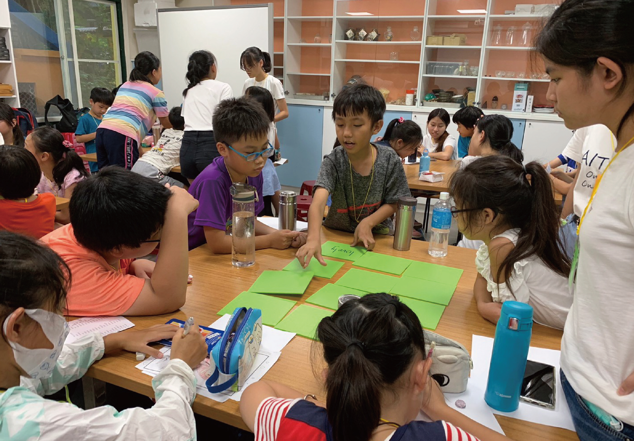 教室內一群小學生圍繞著木桌進行小組活動，桌上擺放著綠色紙張和文具。一名戴眼鏡的男童和其他學生專注地參與課程，講師在旁指導。背景有書櫃和其他學生在活動。