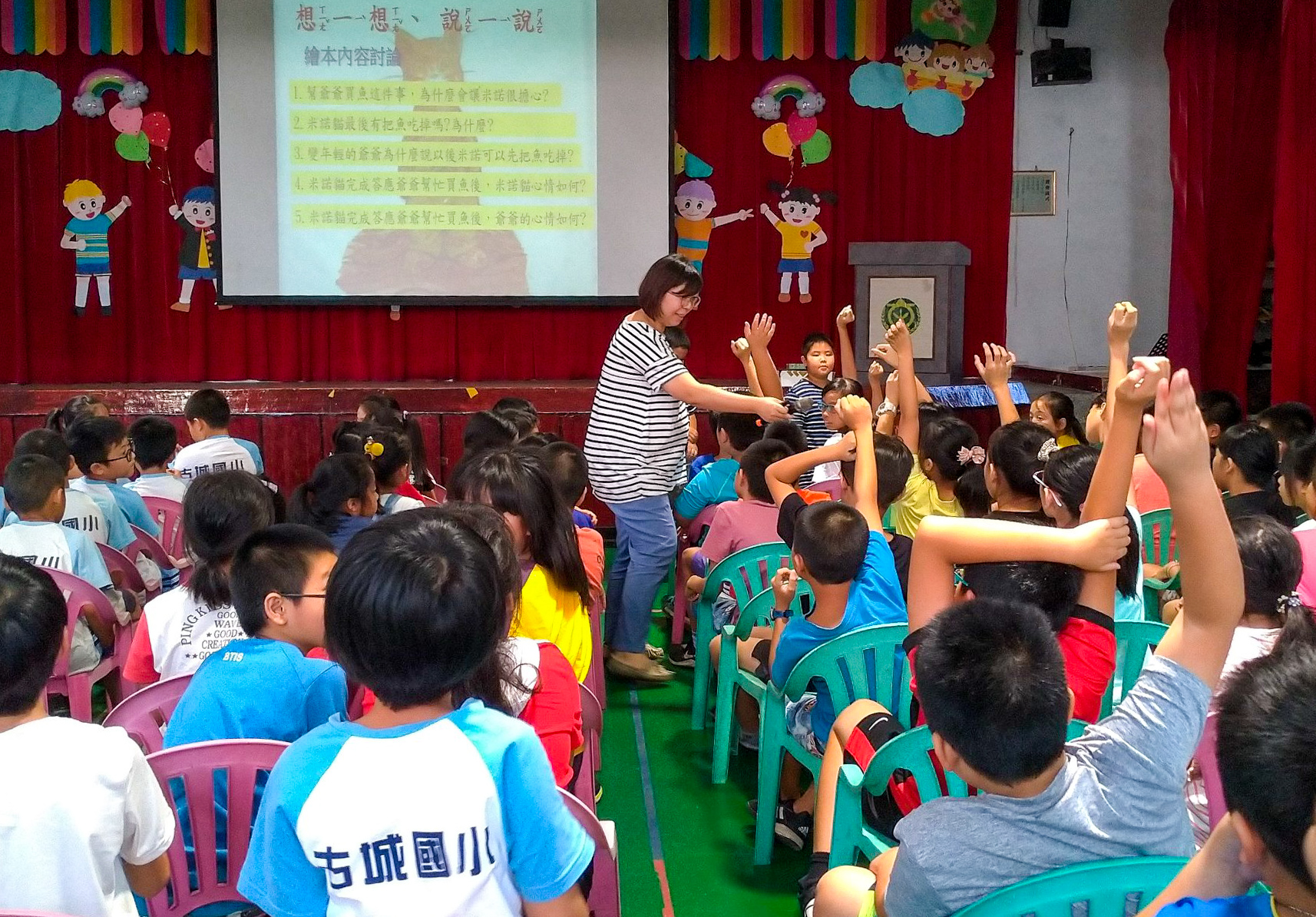 小學教室內，一位女教師在舞台前進行互動教學，眾多穿著校服的學生舉手踴躍參與。背景投影幕上顯示中文教學內容，舞台裝飾有紅色布幕和卡通人物。學生坐在彩色椅子上認真聽講。