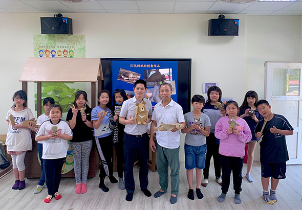 一群小學生和兩位成人在教室內排成一列，面向鏡頭做出勝利手勢。背景有電視螢幕、教室布置和教學用具。這是一場校園活動或教學互動的集體合照。