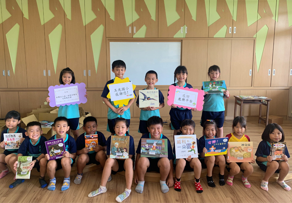一群約十五名小學生在教室內排成兩排，展示他們閱讀的書籍。後排學生站立舉著書籍和學習作品，前排學生坐著展示各類圖書。背景為木製櫃子和白板，呈現溫馨的閱讀推廣活動場景。