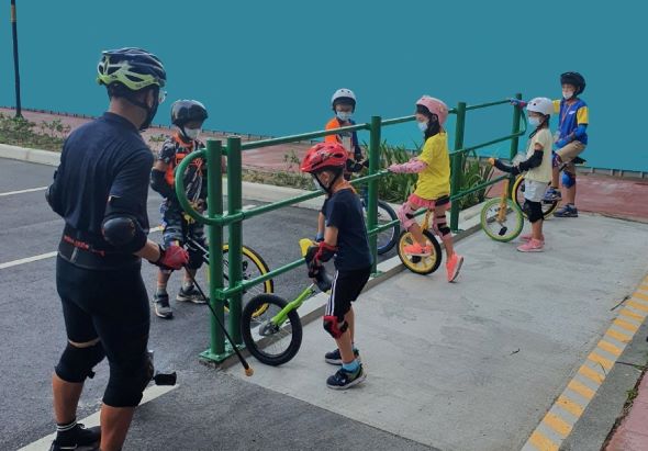 一位成人教練在海邊平台上指導多位兒童騎行平衡車。孩子們穿戴安全頭盔和護具，在綠色欄杆旁排隊練習騎車。清晨陽光下的戶外自行車訓練課程。