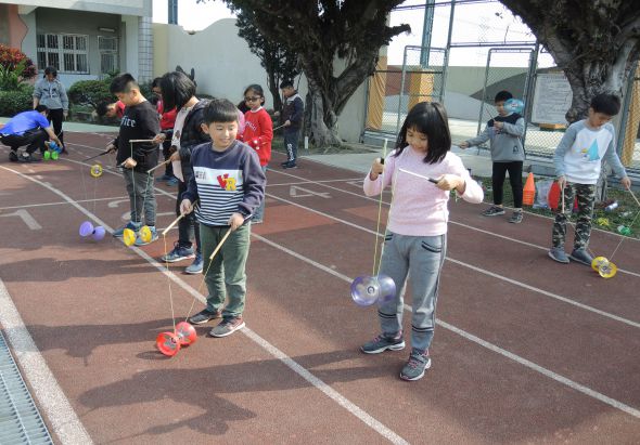 學校操場上，多名兒童正在進行跳繩活動。前景中穿著條紋和粉紅色上衣的兩名學生認真地跳繩，背景有其他學生排隊等候輪流。大樹和校舍圍繞著運動場地，呈現充滿活力的課外活動情景。