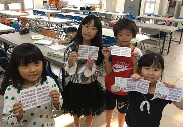 教室內五位小學生展示數學學習成果，每人手持填滿數字的練習紙。孩子們穿著不同顏色衣服，在課桌前排成一列，笑容滿面地展示他們的作業。背景可見多張藍色課桌和教室環境。