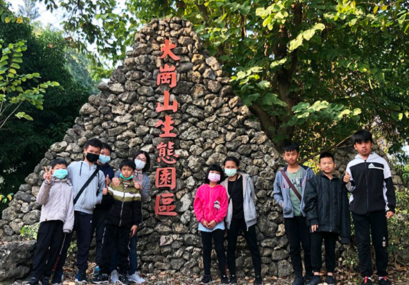 一群學生戴著口罩站在石砌紀念碑前合照，碑上刻著紅色繁體字「岡山美濃園區」。眾人排成一列，背景為茂密的綠色樹木。