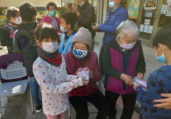 室內活動現場，多位戴口罩的人聚集，中央一位穿著花色上衣的女性和穿著深紅色背心的長者正在互動，周圍有許多民眾參與，背景可見布置和海報，呈現社區聚集或慈善活動的場景。