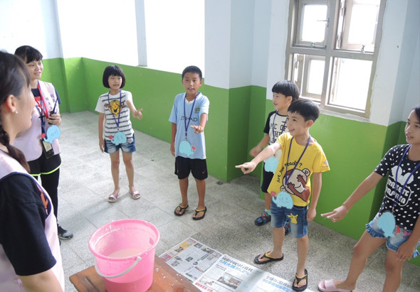 一群小學生在教室裡進行戶外教育活動，站成一圈圍繞著粉紅色水桶和教學資料，老師在左側講解，孩子們穿著各色衣服認真聽講，背景是綠白相間的牆面和窗戶。