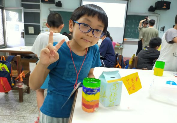 小學教室內，穿著藍色上衣的男童戴著眼鏡，做出勝利手勢面向鏡頭微笑。桌上擺放著彩色堆疊杯和黃色紙張等手工藝品。背景可見其他學生在進行課堂活動，牆上有黑板和教學用具。