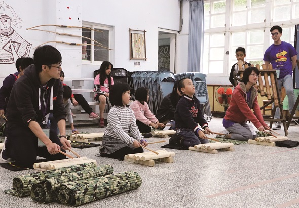 教室內，數名兒童圍坐在地板上，正在進行木製積木或建築材料的手工課程。成人講師在旁指導，牆上掛著教學用圖表。窗邊可見教室環境，其他學生在教室後方活動。