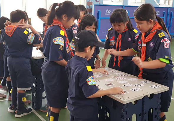 一群穿著童軍制服的小學生在室內聚集於桌前，共同參與一項團隊活動。一名穿著紅背心的領隊正在指導學生們進行桌上遊戲或教學活動，其他學生圍成一圈認真觀看和參與。背景可見藍色的儲物櫃。