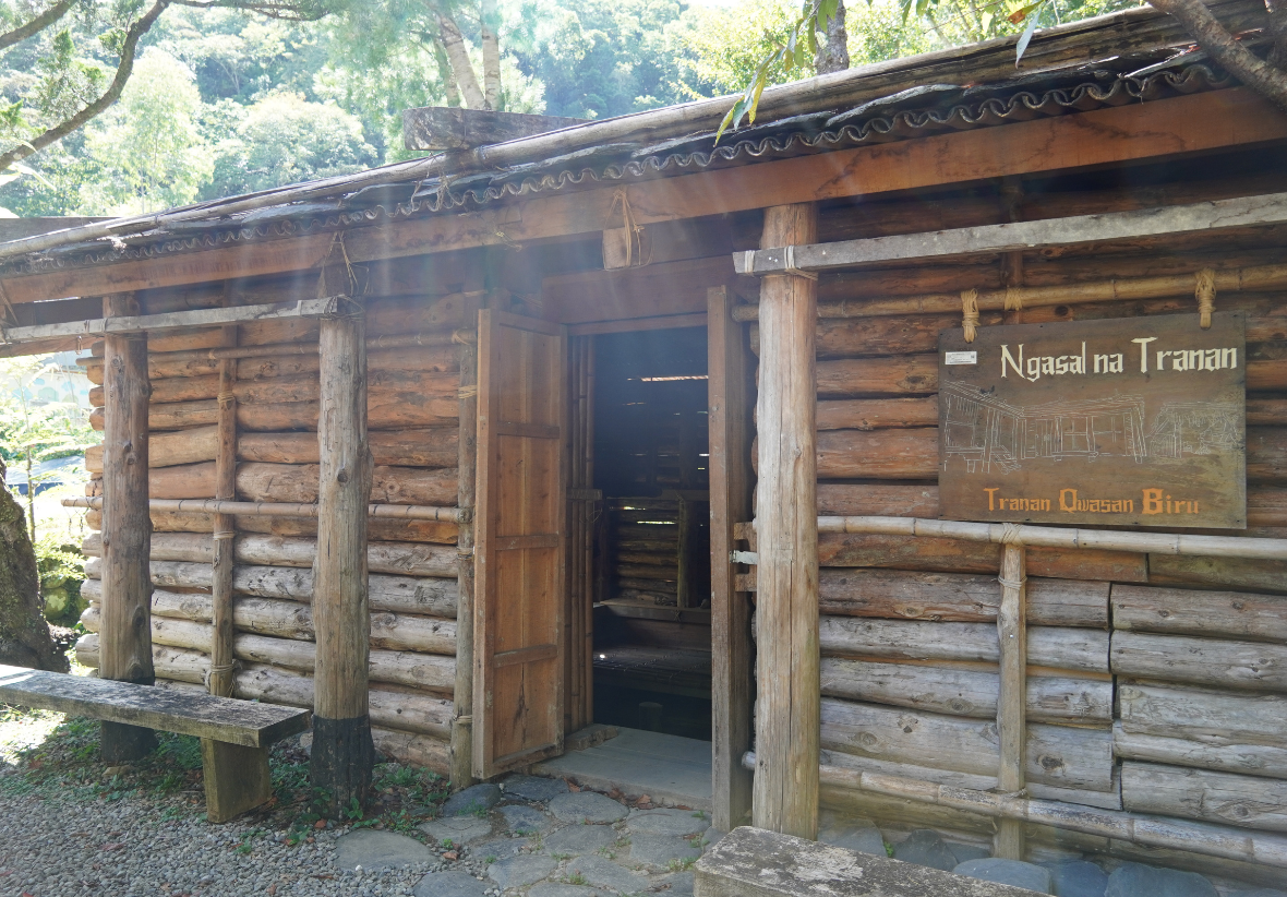 傳統木造建築，具有古樸的橫木牆面和裝飾性屋簷。門口右側掛著黃褐色木製牌匾，上方以白色字體標示「Ngasal na Tranan」，下方標註「Tranan Quisaan Biru」。建築周圍環繞著綠樹，展現原住民傳統建築風格。