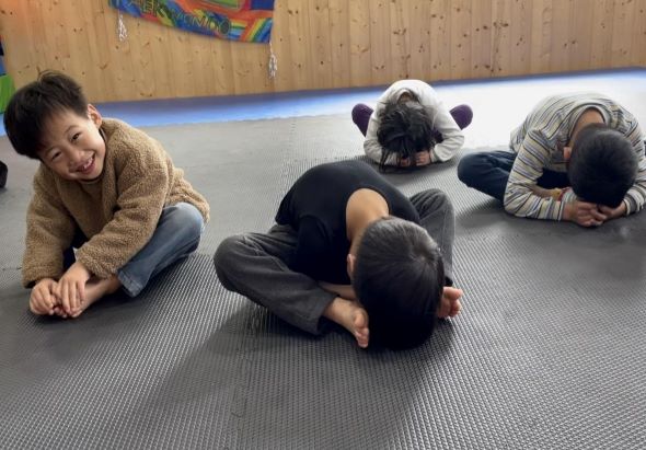 室內體操教室中，一位穿著棕色毛衣的女性教練與四名兒童在灰色墊子上進行瑜伽或伸展運動。其中一名兒童穿著熊貓造型服裝。木質牆面和彩虹旗幟裝飾著教室。
