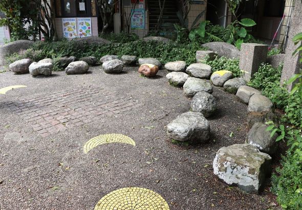 戶外庭園步道，地面鋪設碎石和黃色圓形裝飾磚。兩側排列大小不同的灰色圓形石頭，背景有綠色植物、建築物和告示牌。營造禪意景觀。