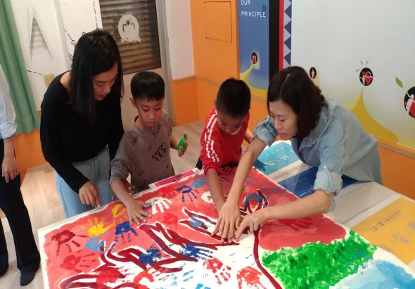 幼兒園教室內，一位女教師與三名幼童圍坐在白色桌邊，共同在紅色大紙張上進行手指繪畫活動。孩子們用多彩顏料創作，牆上張貼著教學海報和彩色裝飾品。