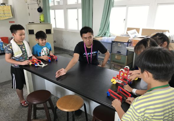 教室內，一位年輕教師在黑色工作台前與五名小學生進行科學或STEM教育活動。學生們正在操作紅色和藍色的機器人積木玩具。教室背景可見綠色窗簾、儲物櫃和教學用具。
