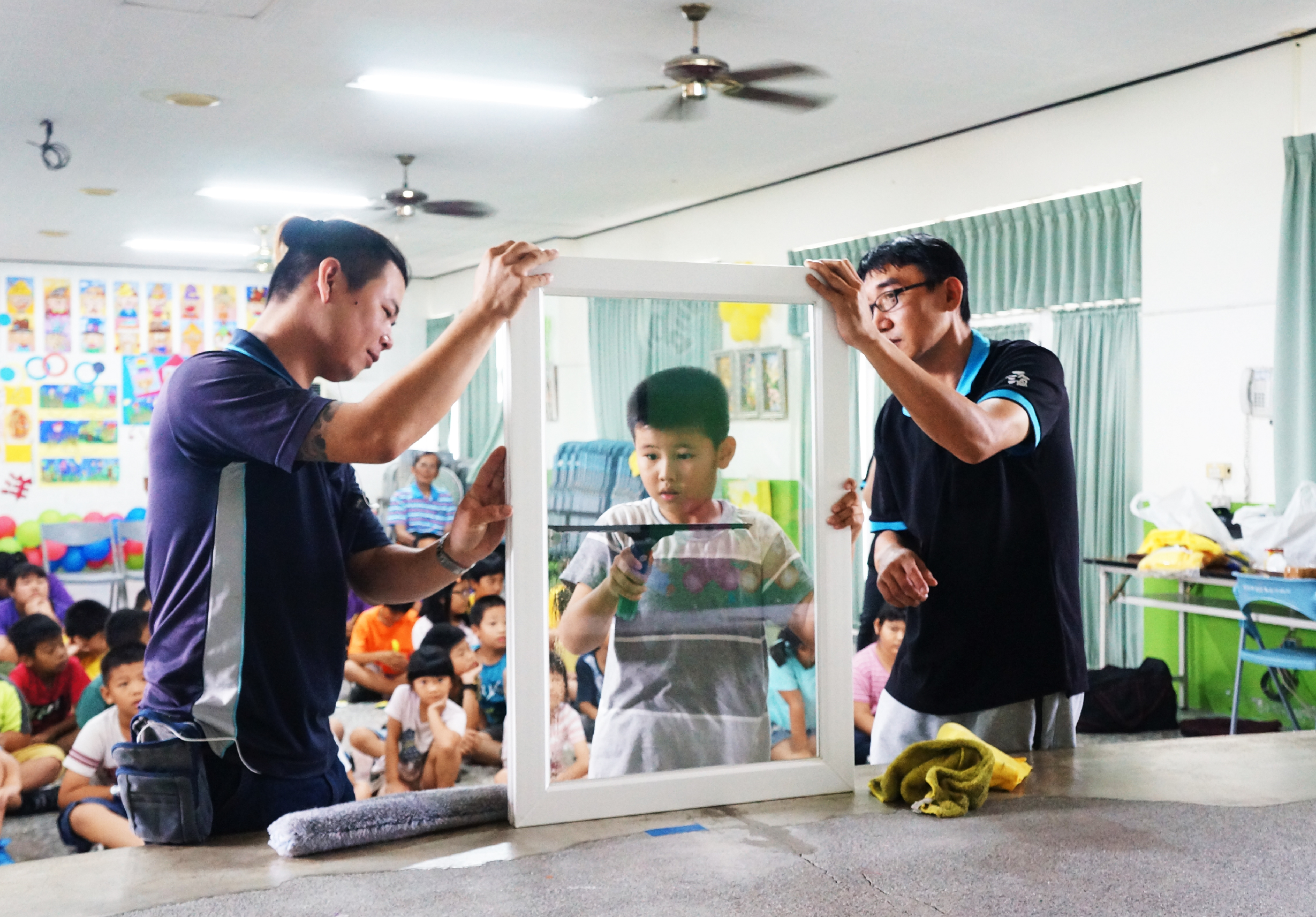 兩位男性講師在幼兒園教室內向坐成一圈的兒童們示範清潔玻璃門窗的方法。教室牆上貼滿彩色作品，多名幼童認真觀看講師的實際操作演示。