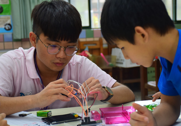 兩名學生在教室內進行電子電路實驗，左邊學生戴著眼鏡，穿著淺紫色上衣，正專注操作電路板和彩色電線，右邊學生穿著藍色上衣，在一旁觀察和協助。桌上擺放著電子元件和實驗工具，背景可見教室設施。