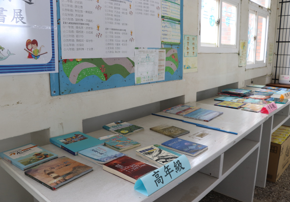 教室內展示桌上擺放多本兒童圖書和教材，牆上貼有課程表、學生作品和彩色教學海報。窗邊放置額外的書籍。整個空間呈現閱讀教學和班級展示的學習環境。