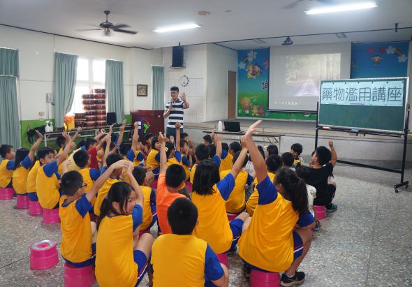 幼兒園教室內，穿著黃藍色制服的小朋友圍坐成圓圈，舉起雙手參與互動課程。講台上的老師正進行教學，身後黑板寫著繁體中文課題，教室配備投影機、風扇及彩色布置。