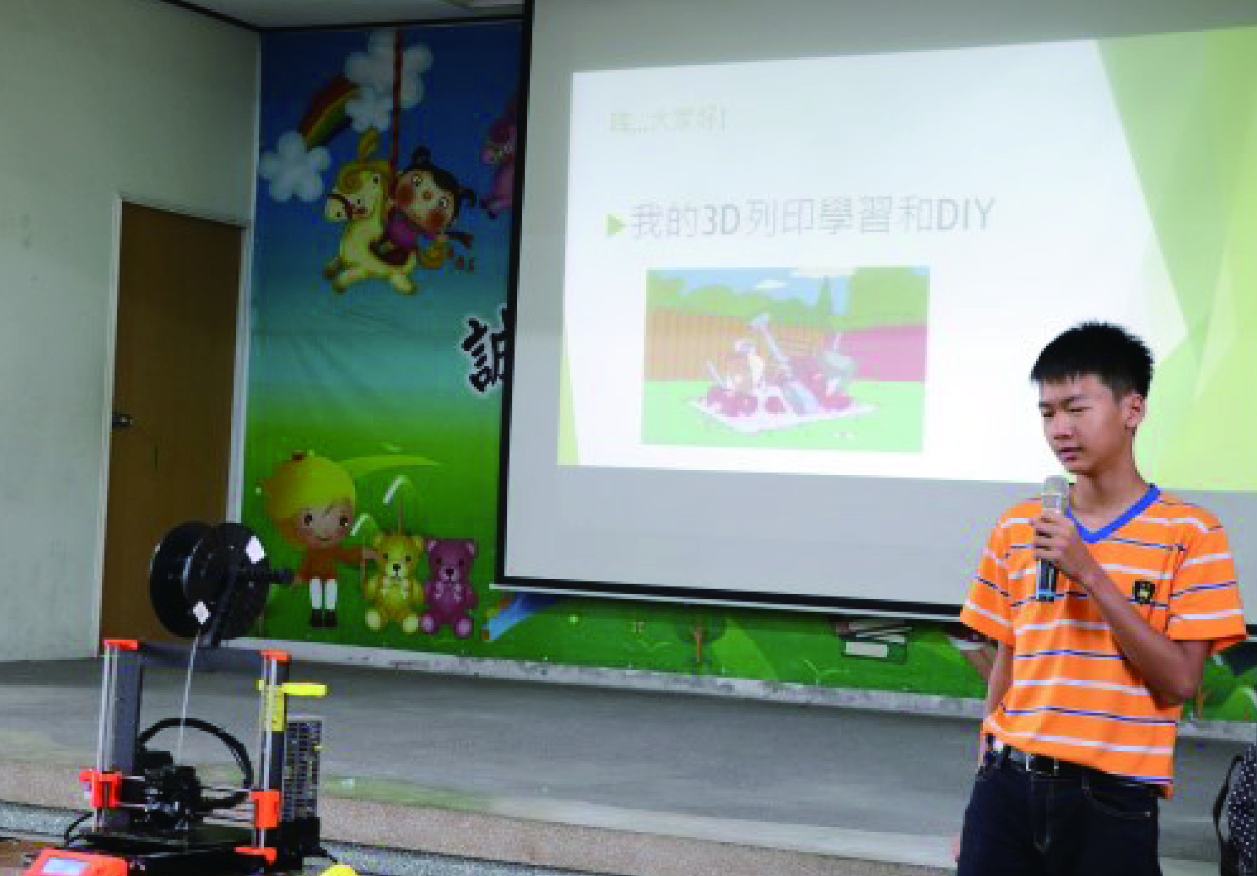 一名穿著橙色條紋上衣的年輕男性在教室進行3D列印與DIY主題的演講。身旁放置橙紅色的3D列印機，投影幕上顯示中文簡報內容與3D列印房屋的示意圖。背後牆面裝飾有卡通人物與綠色背景設計。